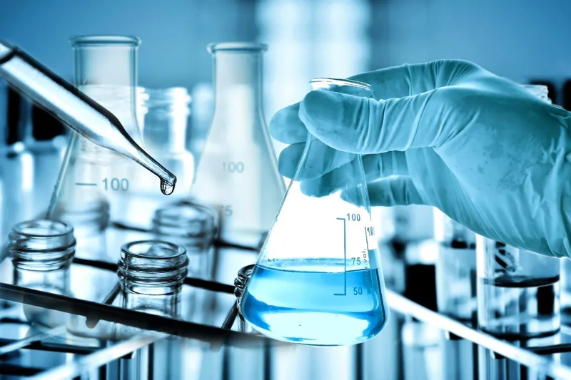 A gloved hand holds a flask with blue liquid about to be mixed, near test tubes and beakers, while a pipette dispenses liquid in a laboratory setting.